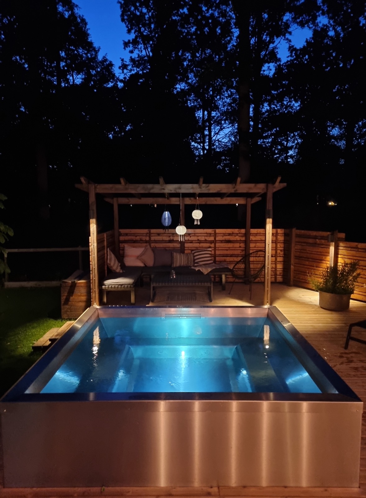 Lit stainless steel spa under a pergola at dusk